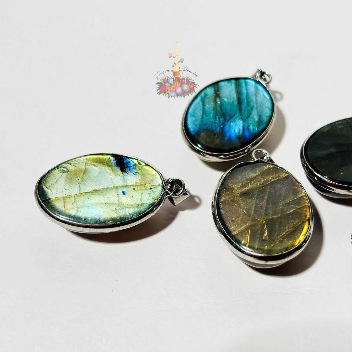 An angled, close-up shot of four oval labradorite pendants in silver settings resting on a crisp white surface. The low angle emphasizes the unique flash (labradorescence) of each gemstone: one in the foreground glows with a bright pale green and yellow flash, another is a deep electric blue and teal hue, a third shows a subtle golden-bronze sheen with natural linear patterns, and a fourth is partially visible on the right.