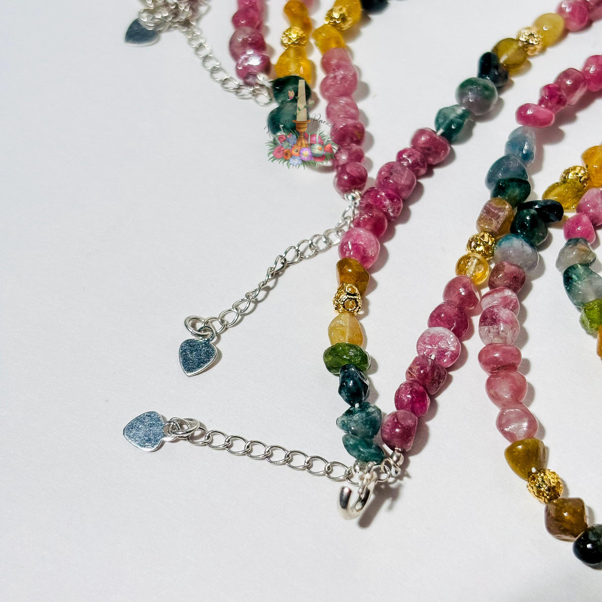 Detail view of layered tourmaline crystal beaded bracelets draped over a white surface, highlighting silver extension chains finished with small, polished heart charms, silver hooks, and textured gold accent beads among pink, green, and yellow gemstones.