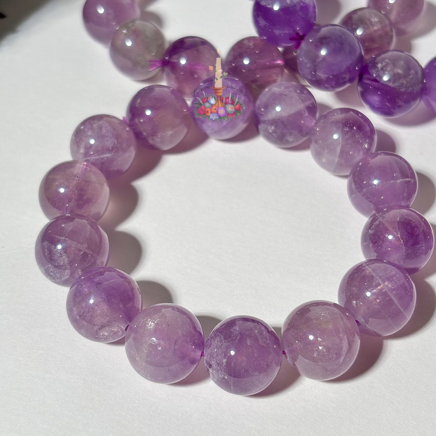 A beaded bracelet made of translucent purple amethyst round beads, featuring a central decorative bead with a colorful floral pattern and small candle details, displayed on a white surface.