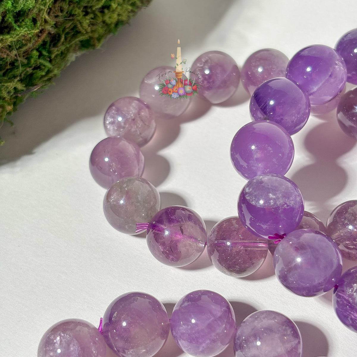 Purple amethyst gemstone round beads arranged in a bracelet or necklace on a white surface, with a small decorative candle graphic near the top center.