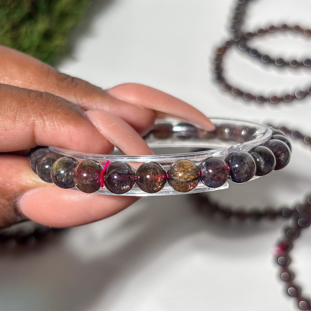 A hand holding a clear plastic bangle holder with a  bracelet made of polished dark brown and purple gemstone beads with gold flecks, with a matching beaded necklace blurred in the background.