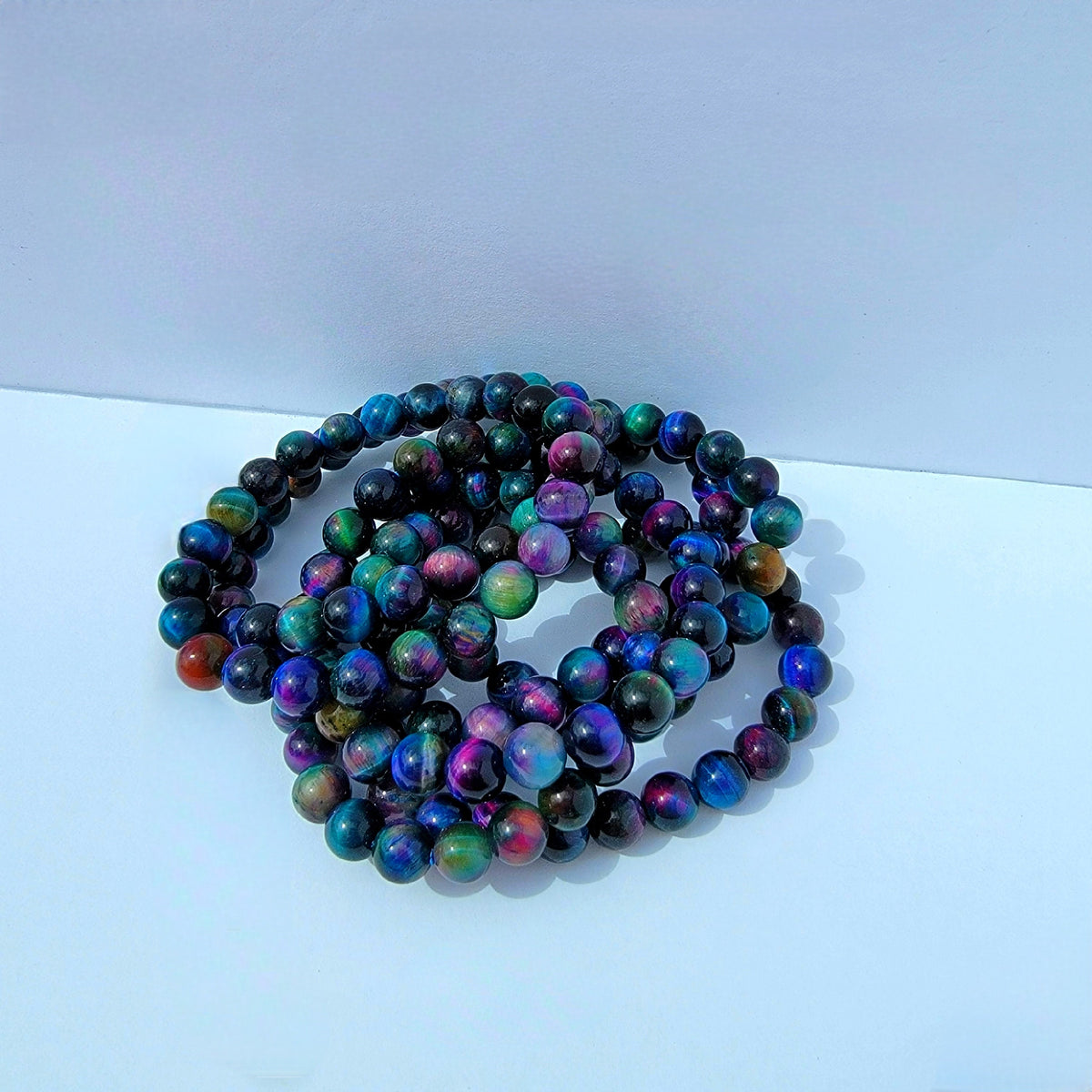 A coiled strand of iridescent round beads with a multicolor, rainbow-like sheen of blue, green, purple, pink, and orange, resting on a plain light gray surface.