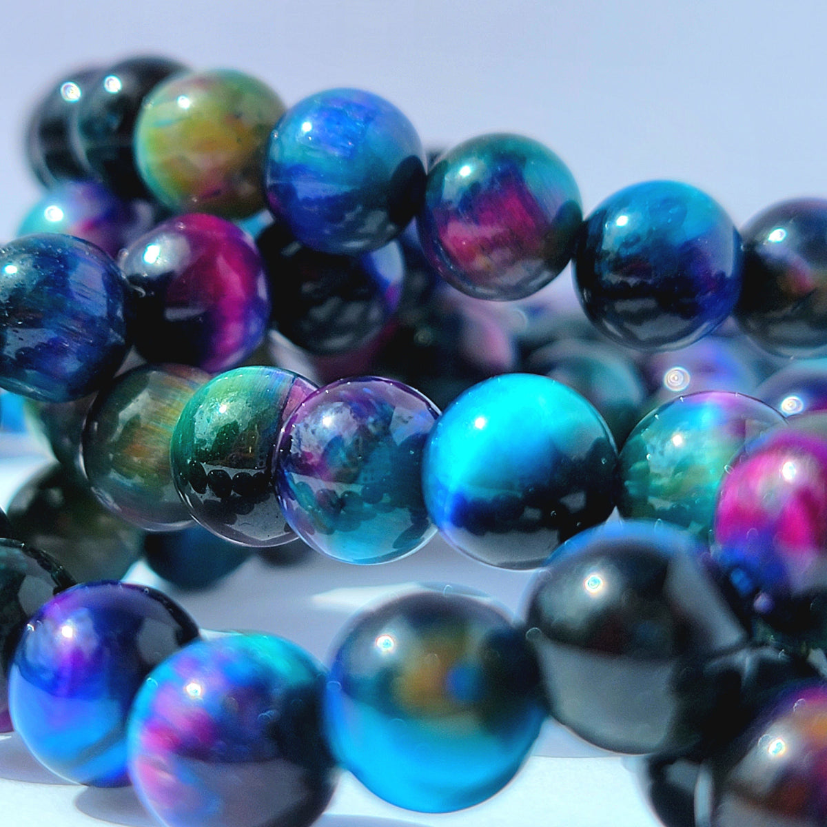 Close-up of glossy round beads with an iridescent galaxy-like finish in shades of blue, purple, teal, and black, arranged in overlapping strands.