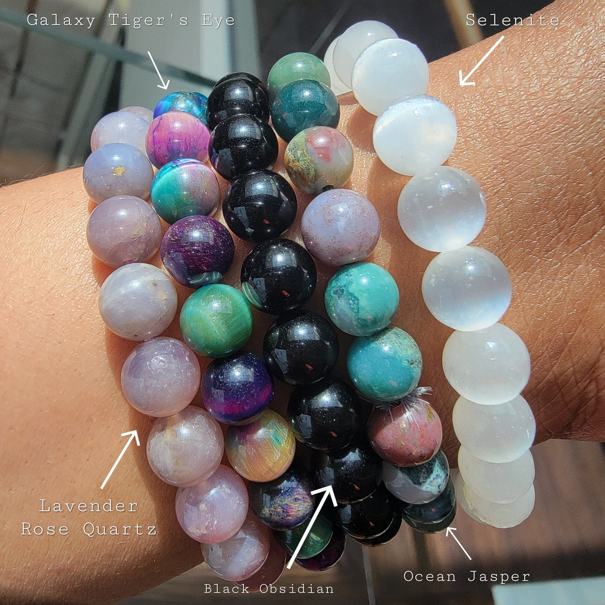 A close-up of a person's wrist wearing multiple beaded bracelets labeled Galaxy Tiger's Eye, Selenite, Lavender Rose Quartz, Black Obsidian, and Ocean Jasper.