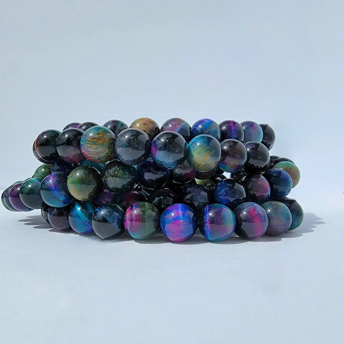 A stack of iridescent beaded bracelets made of round dark beads that shift between blue, purple, green, and pink, photographed on a clean light gray background.
