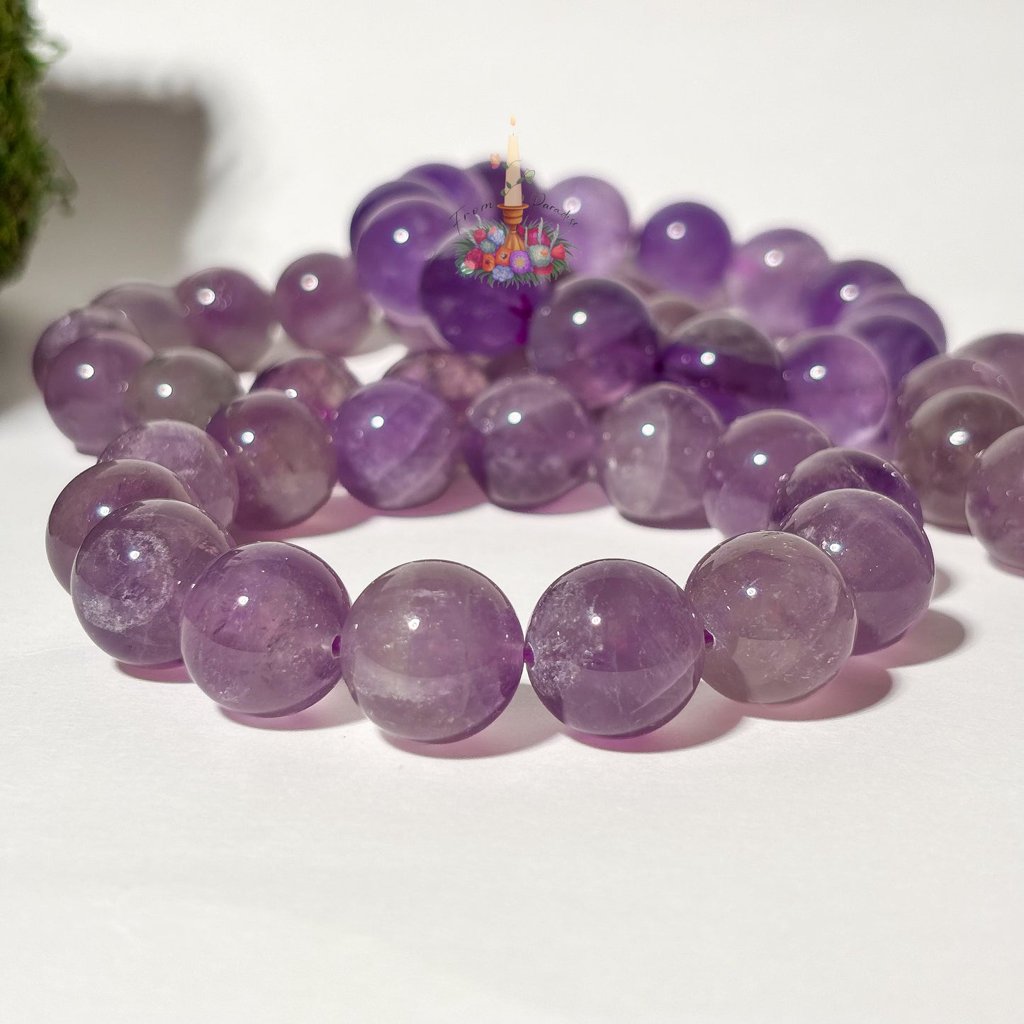 A beaded bracelet made of smooth, polished purple amethyst round beads, arranged in a circle on a white surface with a small decorative floral element in the background.