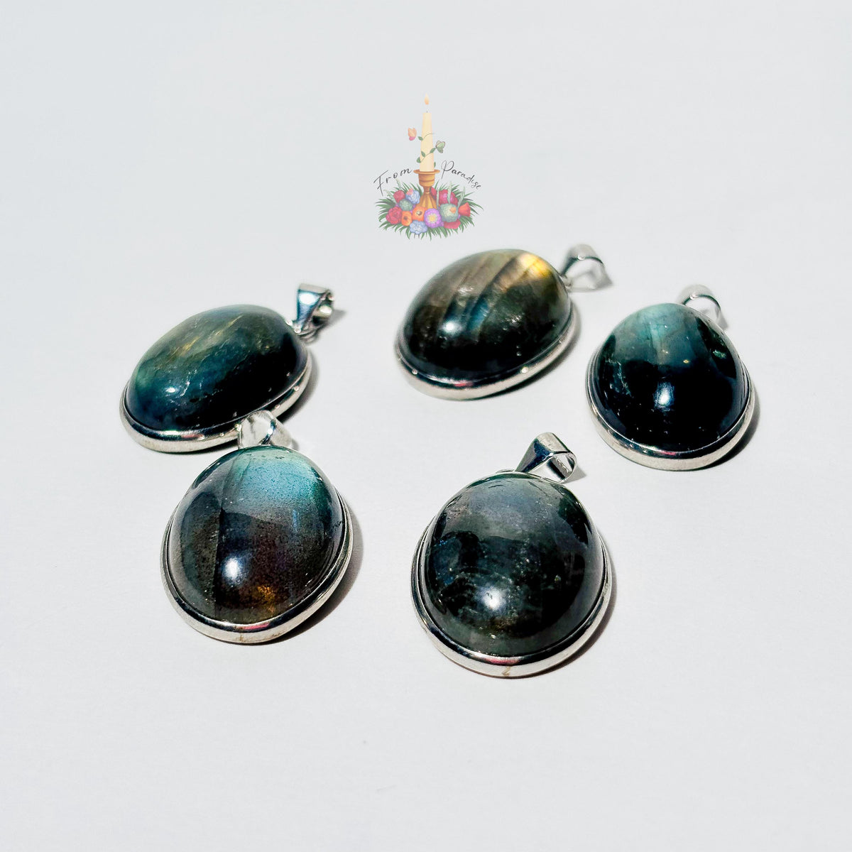 Five oval labradorite crystal pendants set in silver bezels are arranged on a plain white surface. Each pendant showcases a unique natural flash of color, ranging from bright yellow and blue to deep green and grey. A small patch of green moss is visible in the top right corner, and a small decorative logo with a candle and flowers sits in the upper center.