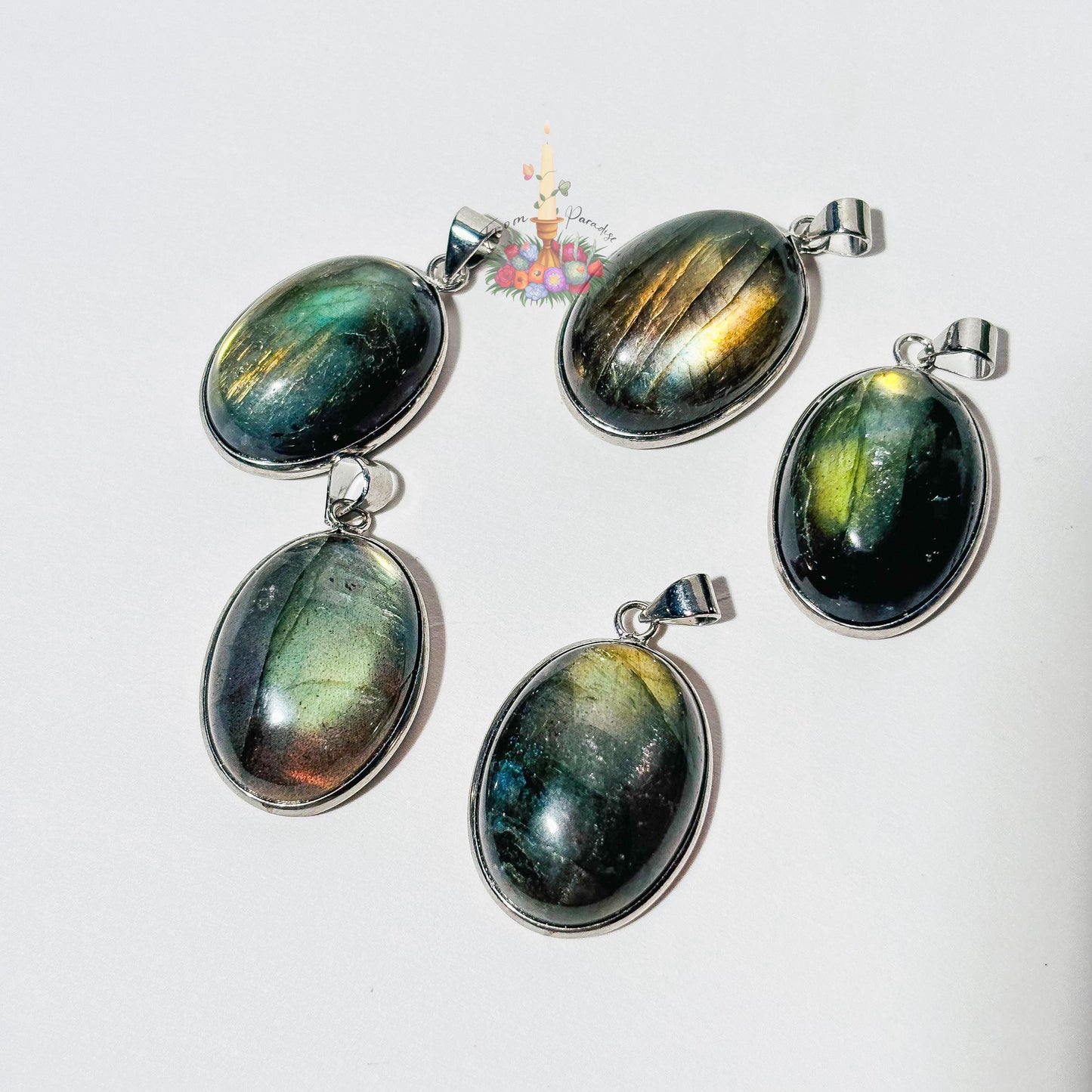 Five oval labradorite crystal pendants set in silver bezels are arranged on a plain white surface. Each pendant showcases a unique natural flash of color, ranging from bright yellow and blue to deep green and grey. A small patch of green moss is visible in the top right corner, and a small decorative logo with a candle and flowers sits in the upper center.