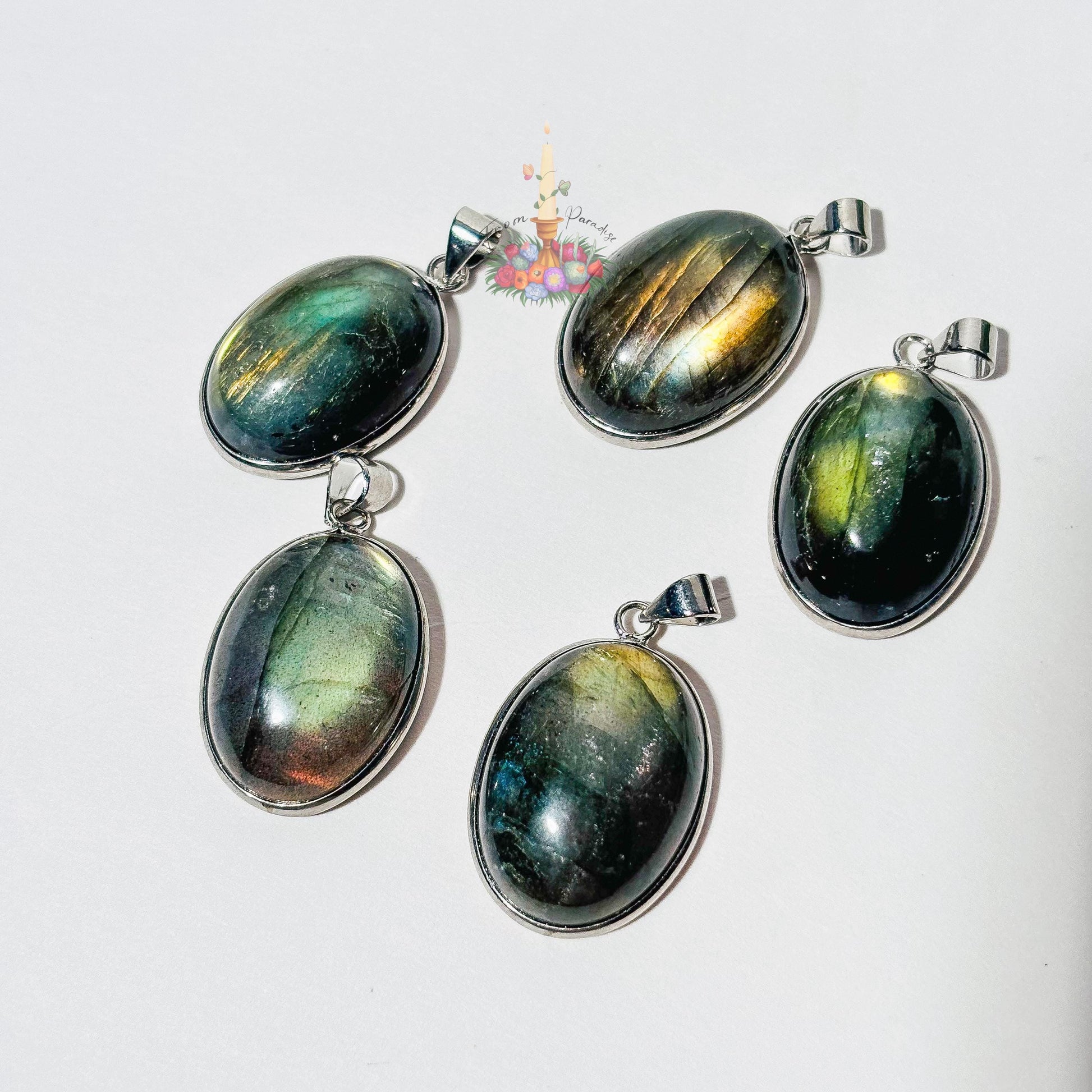 Five oval labradorite crystal pendants set in silver bezels are arranged on a plain white surface. Each pendant showcases a unique natural flash of color, ranging from bright yellow and blue to deep green and grey. A small patch of green moss is visible in the top right corner, and a small decorative logo with a candle and flowers sits in the upper center.