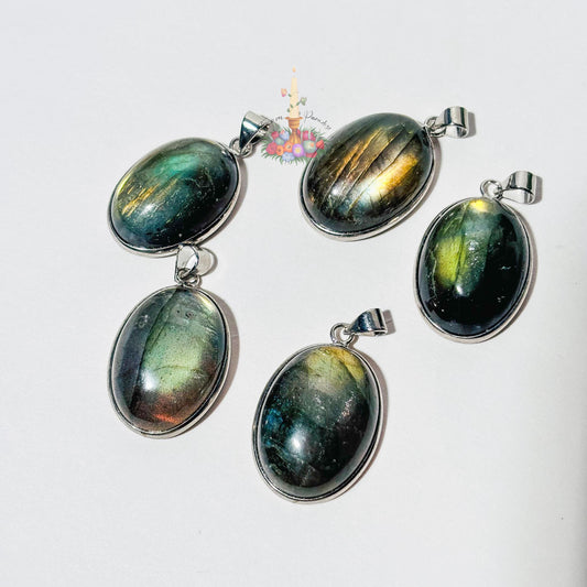 Five oval labradorite crystal pendants set in silver bezels are arranged on a plain white surface. Each pendant showcases a unique natural flash of color, ranging from bright yellow and blue to deep green and grey. A small patch of green moss is visible in the top right corner, and a small decorative logo with a candle and flowers sits in the upper center.