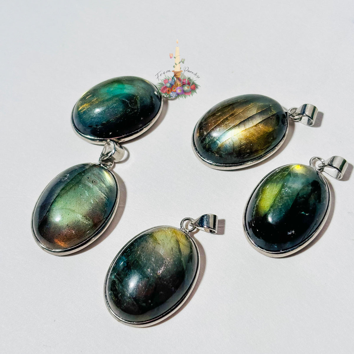 Five oval labradorite crystal pendants set in silver bezels are arranged on a plain white surface. Each pendant showcases a unique natural flash of color, ranging from bright yellow and blue to deep green and grey. A small patch of green moss is visible in the top right corner, and a small decorative logo with a candle and flowers sits in the upper center.