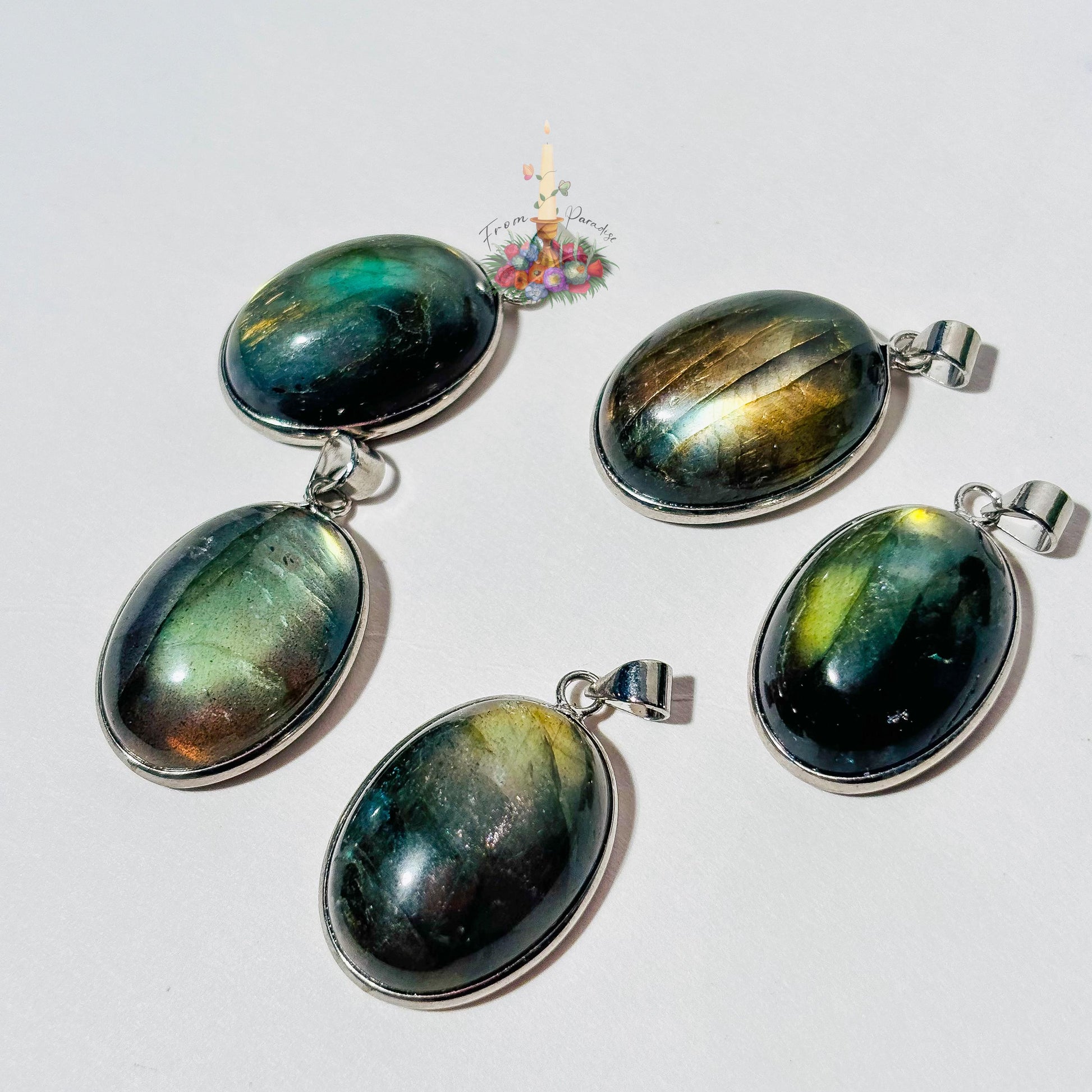 Five oval labradorite crystal pendants set in silver bezels are arranged on a plain white surface. Each pendant showcases a unique natural flash of color, ranging from bright yellow and blue to deep green and grey. A small patch of green moss is visible in the top right corner, and a small decorative logo with a candle and flowers sits in the upper center.