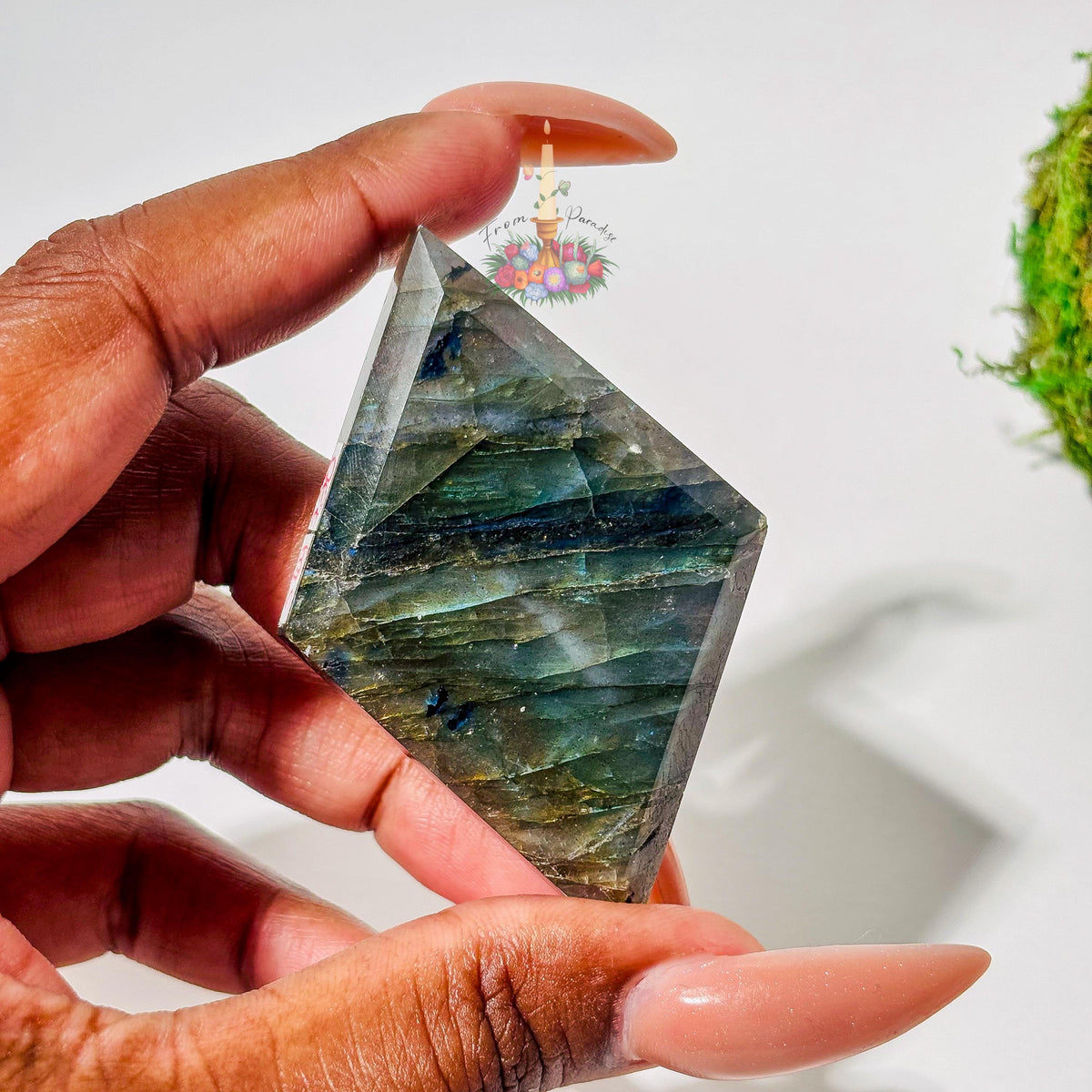 A person's hand with long, neutral-toned nails holds a faceted, diamond-shaped labradorite gemstone. The stone displays deep grayish-green tones with delicate purple and pink chatoyancy across the center and a pop of warm gold flash on the right edge against a white background.