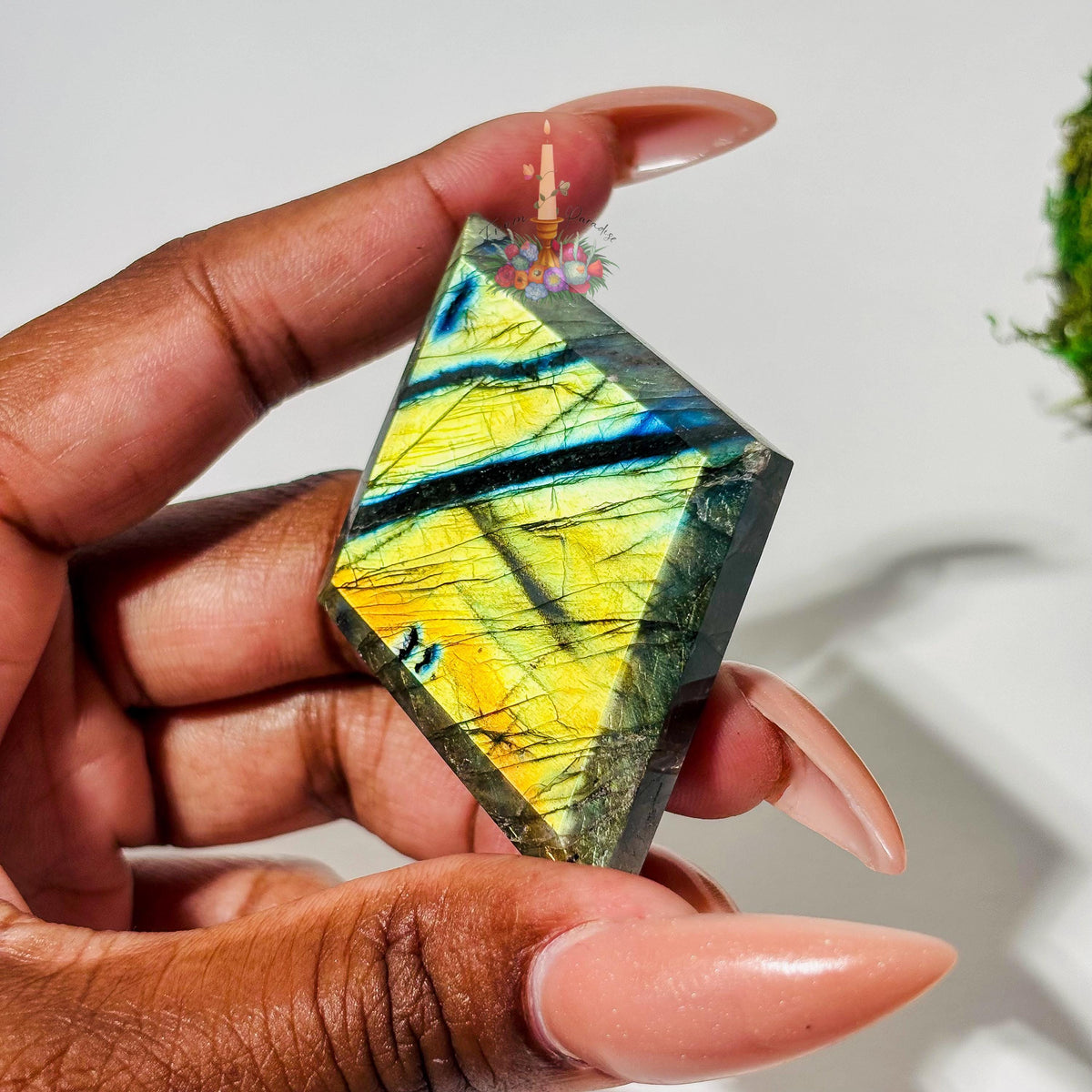 A person's hand with long, neutral-toned nails holds a faceted, diamond-shaped labradorite gemstone up to the camera. The stone features intense flash with striking shades of bright yellow, gold, and teal blue accents highlighting its natural internal fractures against a neutral white background.