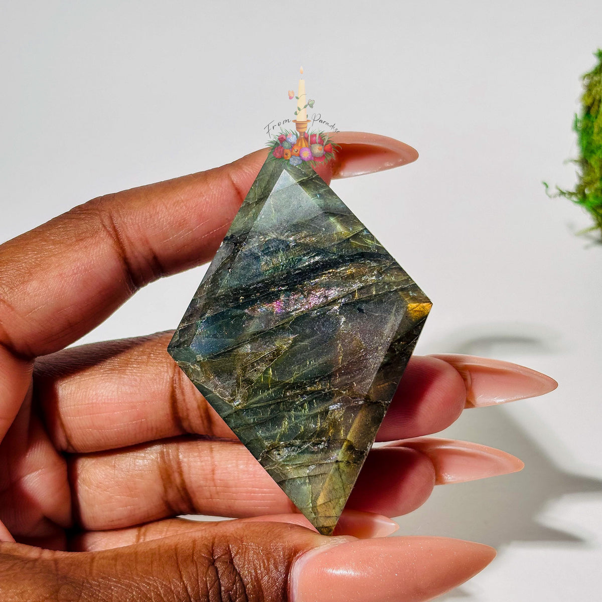 A person's hand with long, neutral-toned nails holds a faceted, diamond-shaped labradorite gemstone. The stone displays deep grayish-green tones with delicate purple and pink chatoyancy across the center and a pop of warm gold flash on the right edge against a white background.