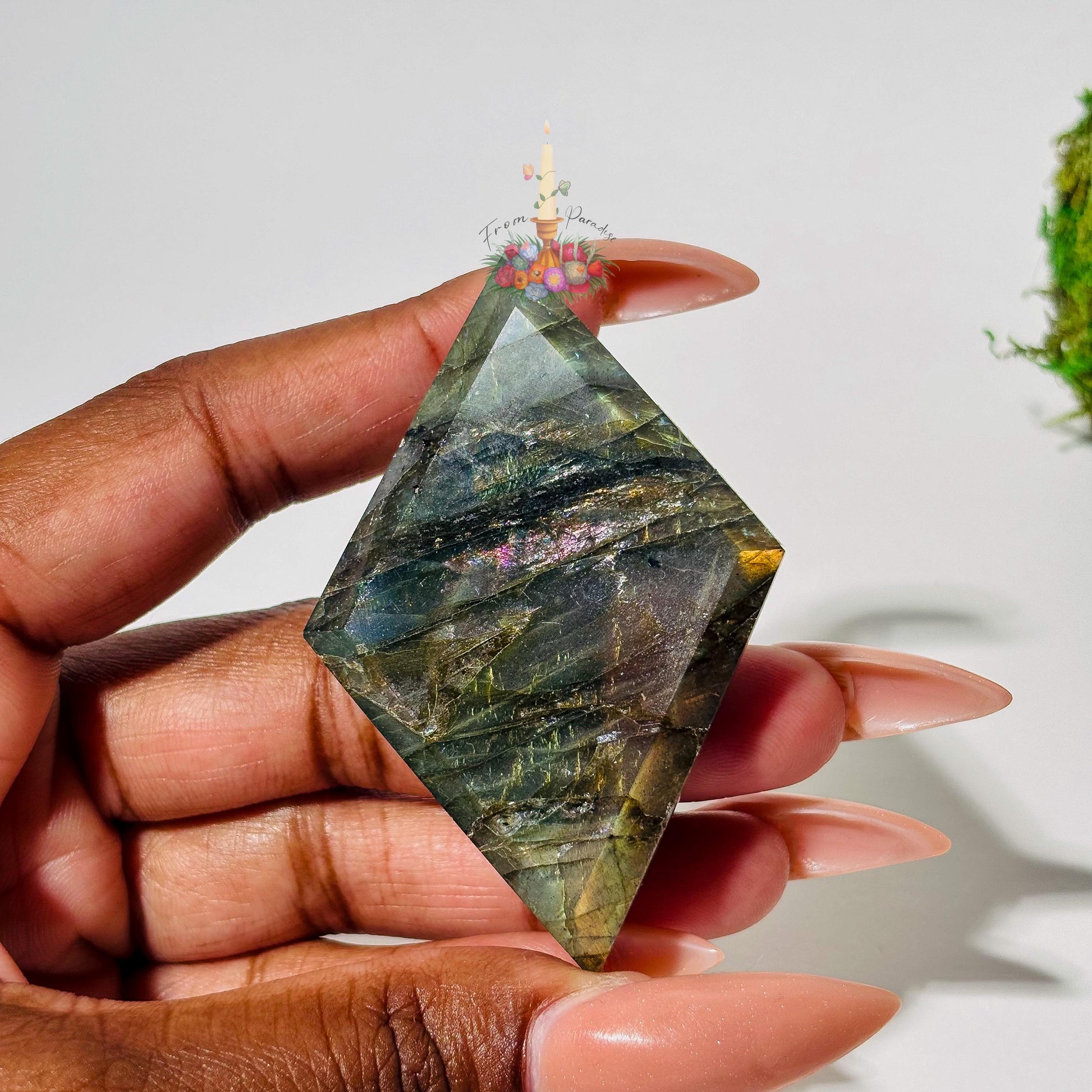 A person's hand with long, neutral-toned nails holds a faceted, diamond-shaped labradorite gemstone. The stone displays deep grayish-green tones with delicate purple and pink chatoyancy across the center and a pop of warm gold flash on the right edge against a white background.