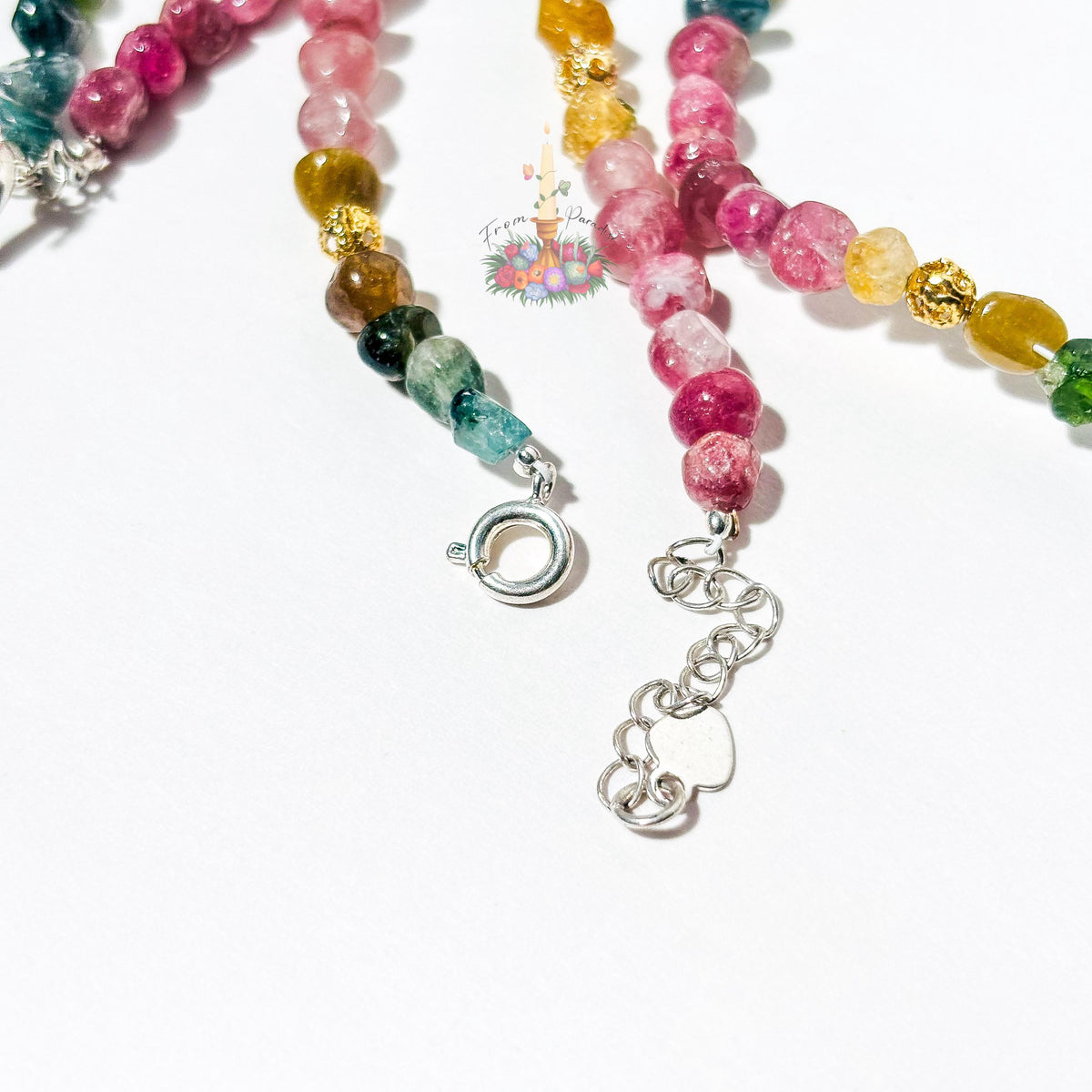 Macro photograph of handcrafted tourmaline gemstone bracelets on a white surface, focusing on a silver spring ring clasp connected to a silver extension chain with a heart charm. Pink, teal, and amber crystal beads are accented with textured gold spacers.