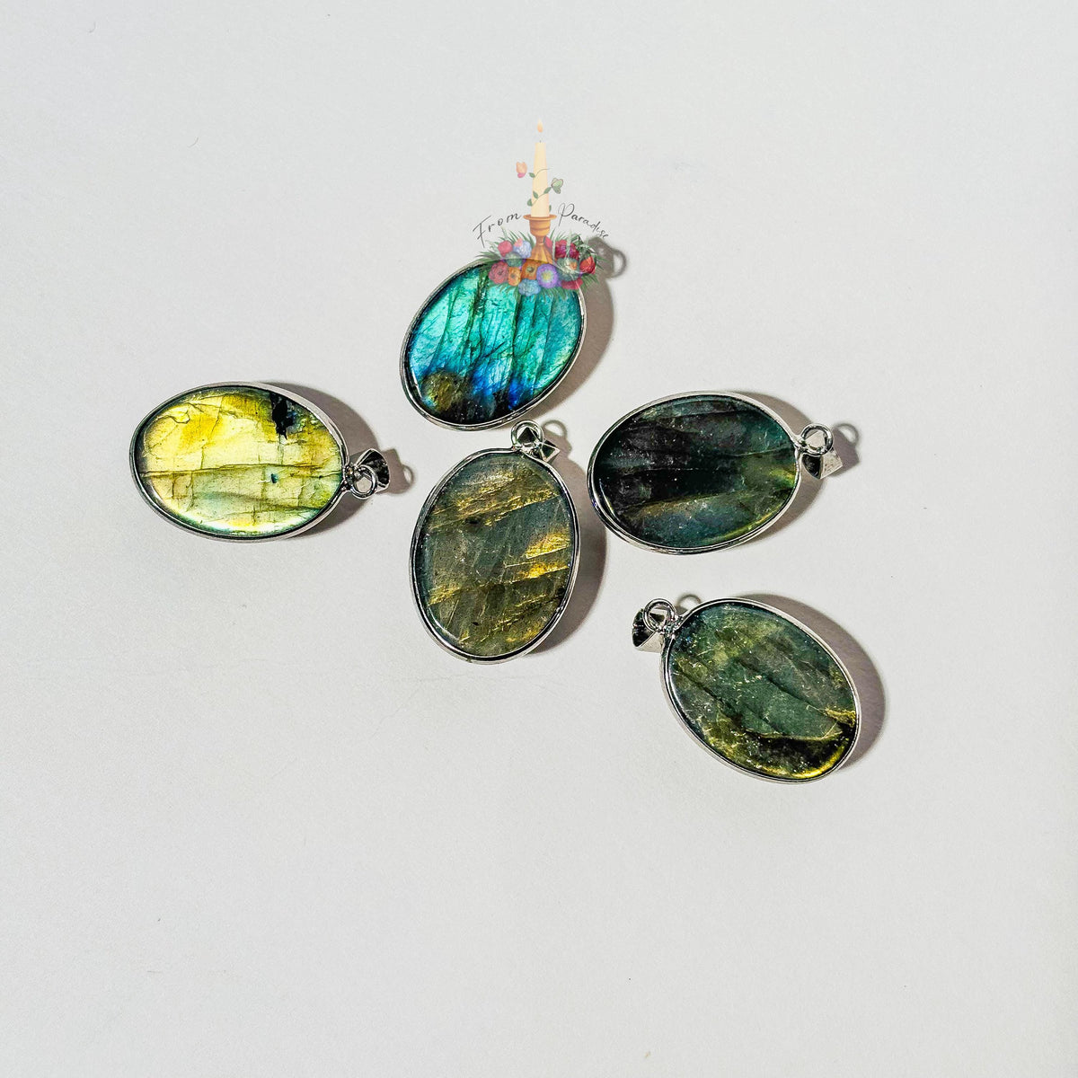 Five oval labradorite crystal pendants set in silver bezels are arranged on a plain white surface. Each pendant showcases a unique natural flash of color, ranging from bright yellow and blue to deep green and grey. A small patch of green moss is visible in the top right corner, and a small decorative logo with a candle and flowers sits in the upper center.