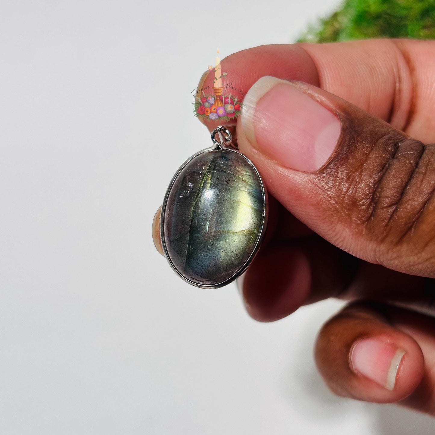 A close-up shot of a person with a medium-to-dark complexion holding a single oval labradorite pendant between their thumb and forefinger to show its scale. The cabochon crystal is set in a silver bezel and displays a moody, metallic flash of golden-yellow and soft blue light across its smooth surface. The background is a clean, minimalist white, with a faint hint of green moss in the upper right and a small candle logo positioned just above the pendant