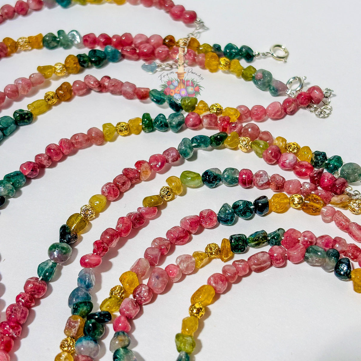 Overhead shot of several colorful tourmaline gemstone bracelets laid out in parallel wave patterns on a white background. The tumbled gemstone beads showcase vibrant shades of watermelon pink, yellow-amber, deep green, and teal, separated at regular intervals by textured gold accent beads with silver clasp hardware visible near the top.