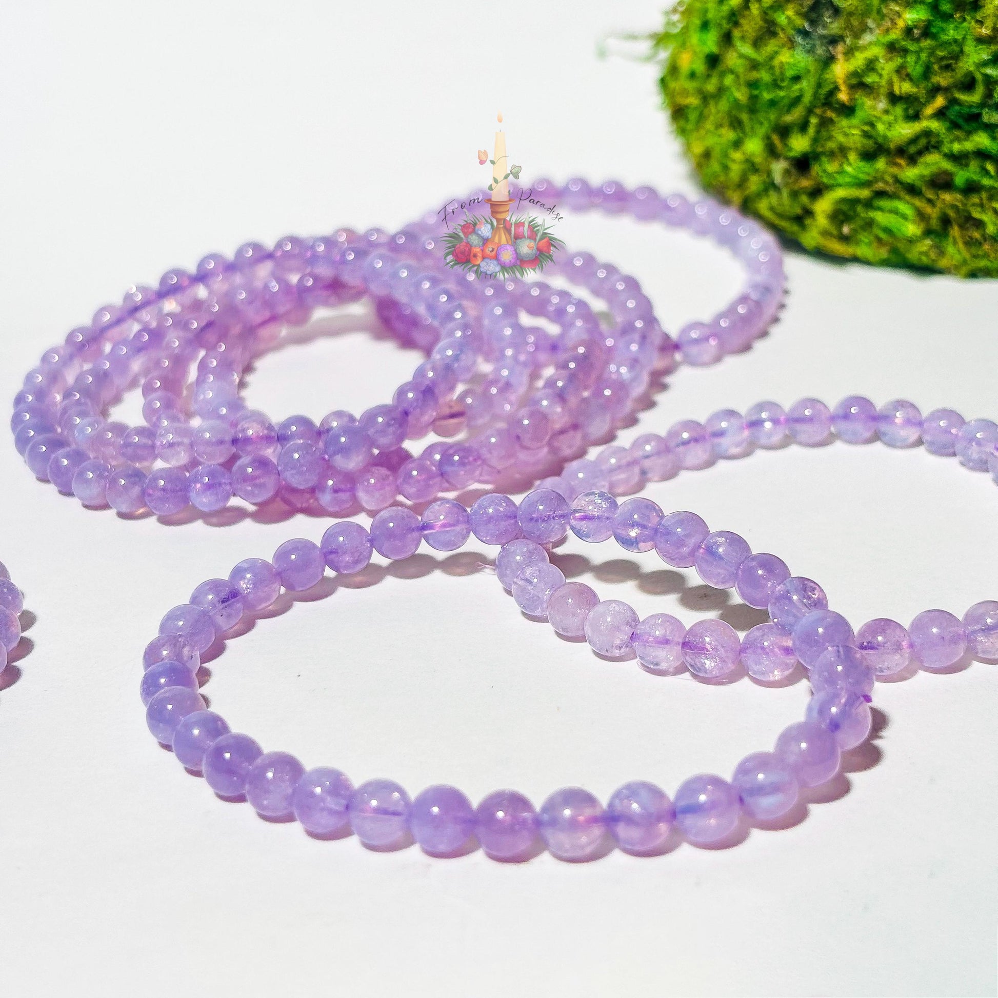 A beaded stretch bracelet made of translucent, pastel lavender moon quartz polished round beads rests on a smooth white surface. In the background, a second matching beaded bracelet is softly blurred, alongside vibrant green moss and a floral candle logo.