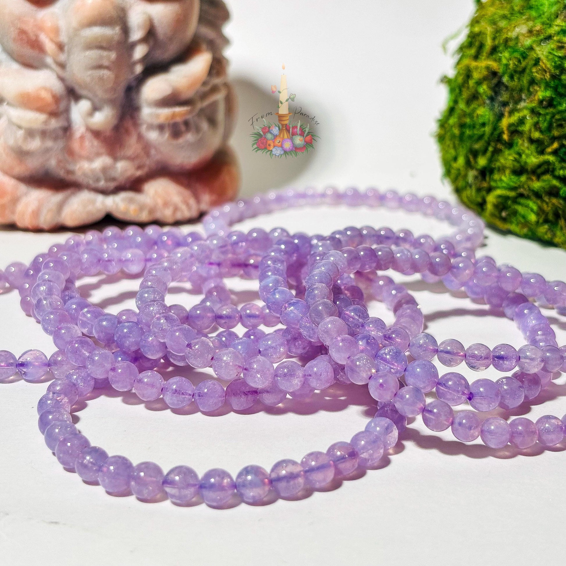 A cluster of round Lavender Moon Quartz beaded stretch bracelets piled together on a plain white surface. A carved stone statue sits out of focus in the background on the left, with vibrant green moss on the right and a floral candle logo centered above the bracelets.