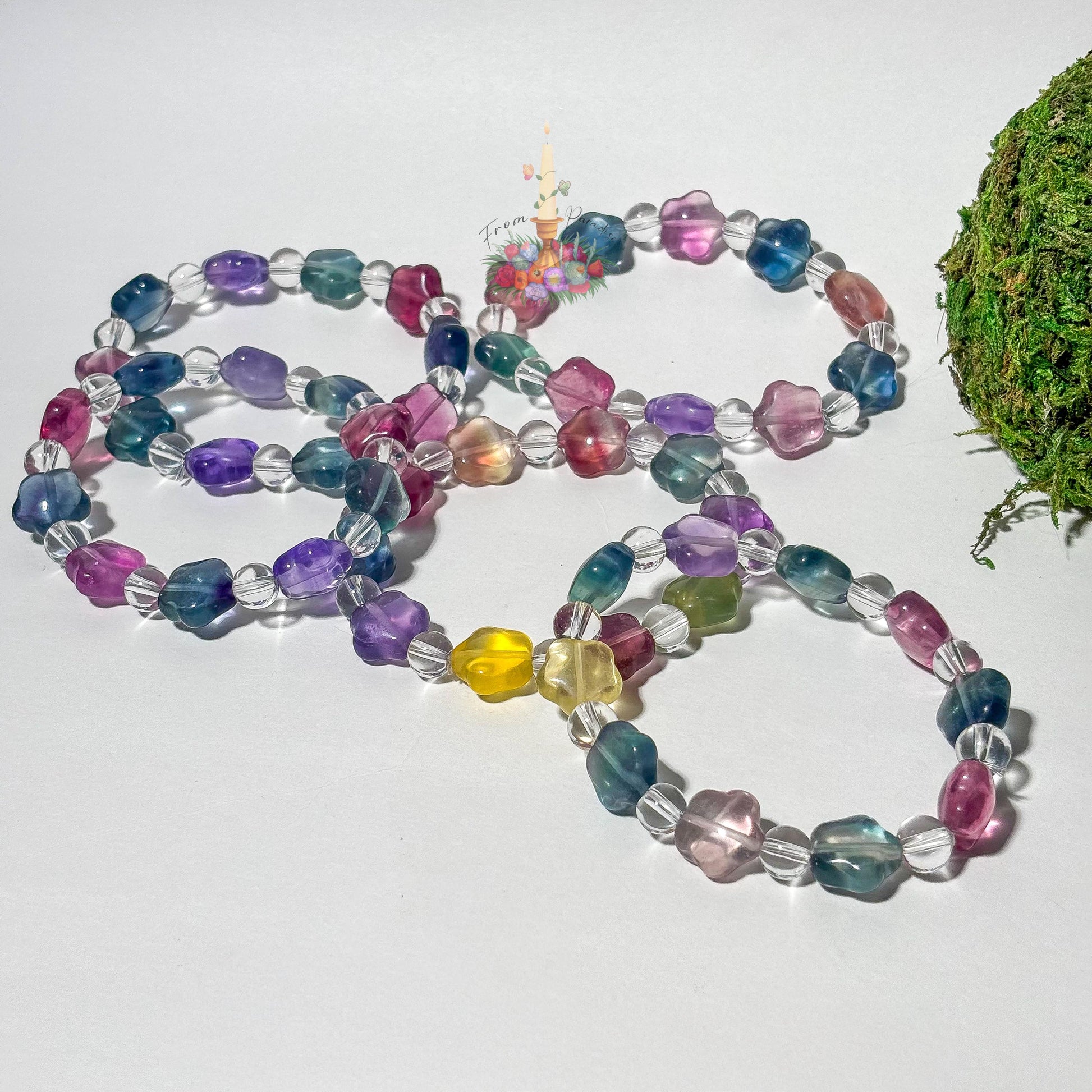 A collection of beaded stretch bracelets featuring star-shaped fluorite beads in translucent shades of purple, teal, magenta, and yellow, alternating with small, round clear quartz beads. The bracelets are arranged on a clean white surface with a patch of green moss on the right and a central floral candle logo.
