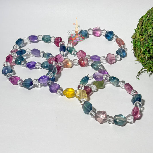 A collection of beaded stretch bracelets featuring star-shaped fluorite beads in translucent shades of purple, teal, magenta, and yellow, alternating with small, round clear quartz beads. The bracelets are arranged on a clean white surface with a patch of green moss on the right and a central floral candle logo.