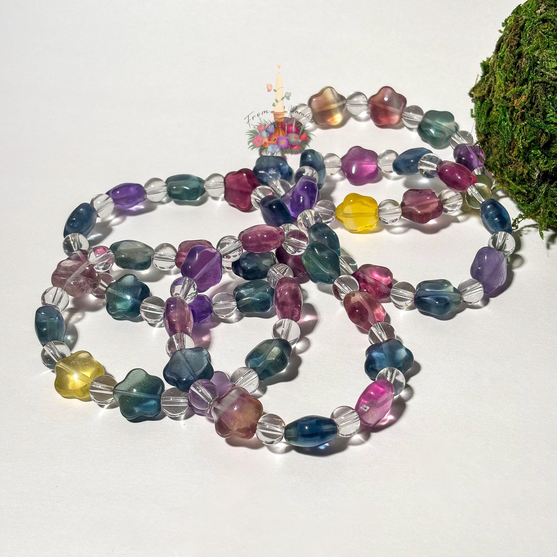A collection of beaded stretch bracelets featuring star-shaped fluorite beads in translucent shades of purple, teal, magenta, and yellow, alternating with small, round clear quartz beads. The bracelets are arranged on a clean white surface with a patch of green moss on the right and a central floral candle logo.