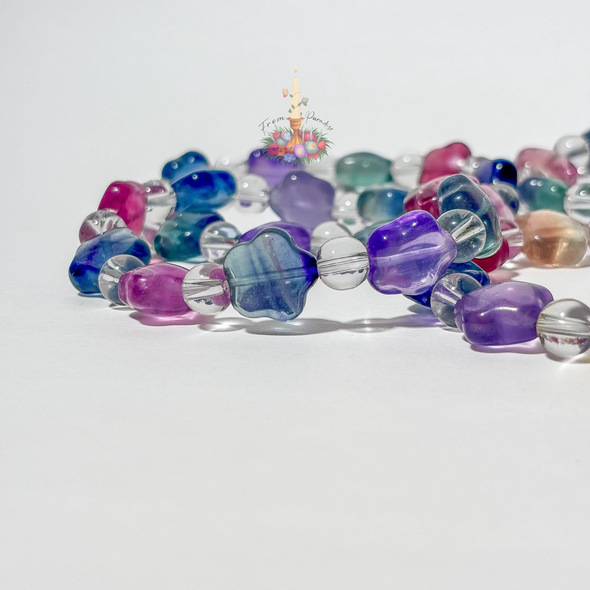 A close-up view of several beaded stretch bracelets on a white surface, with the foreground focusing on translucent, star-shaped fluorite beads in vibrant shades of magenta, blue, and yellow-purple color-zoning, separated by round clear quartz spacer beads. Additional matching fluorite and clear quartz bracelets lie softly out of focus in the background beneath a small floral candle logo.