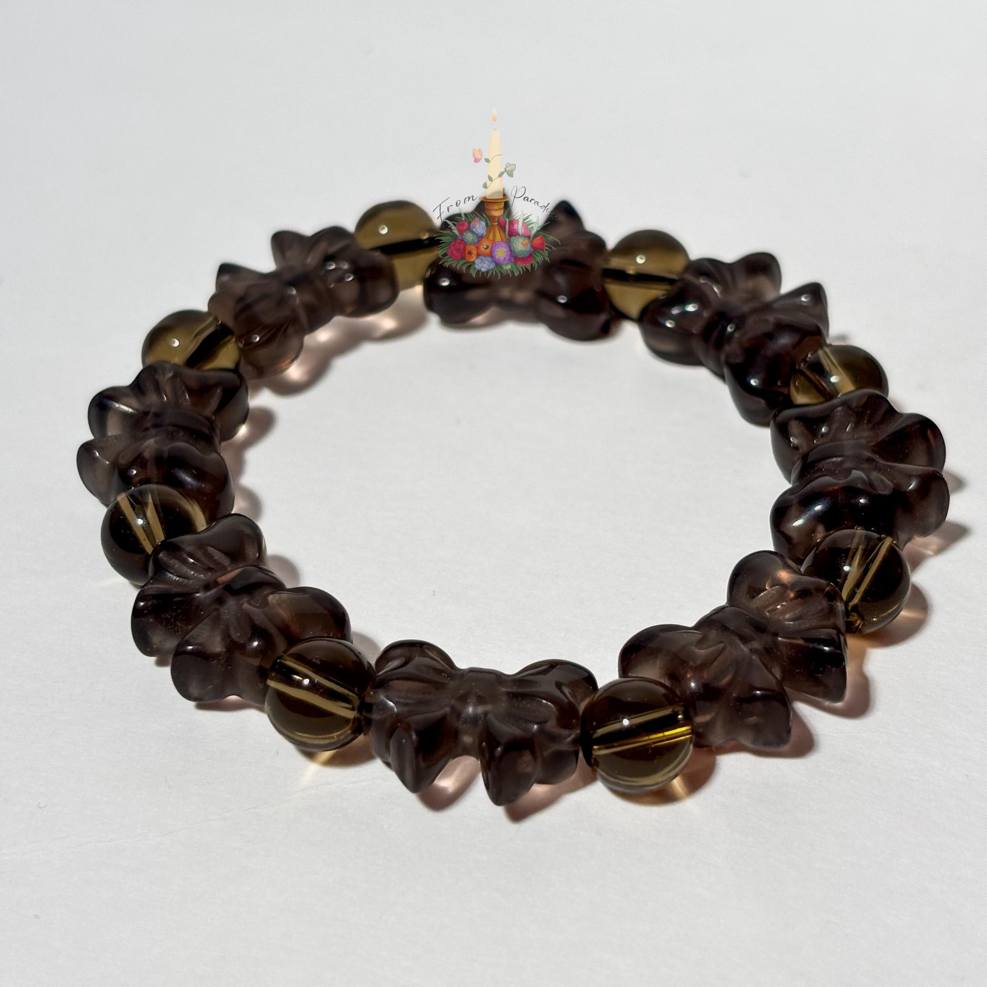 A single beaded stretch bracelet crafted from translucent ice obsidian, arranged in a circular loop against a clean white surface. The bracelet features carved ribbon-bow ice obsidian beads alternating with round spacer beads in the same smoky, translucent brown tone. A small floral candle logo sits near the top center of the loop, with a touch of green moss visible along the right edge.