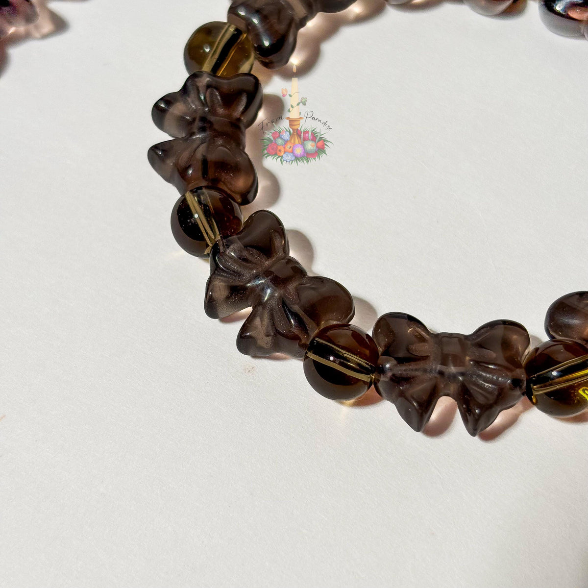 A single beaded stretch bracelet crafted from translucent ice obsidian, arranged in a circular loop against a clean white surface. The bracelet features carved ribbon-bow ice obsidian beads alternating with round spacer beads in the same smoky, translucent brown tone. A small floral candle logo sits near the top center of the loop, with a touch of green moss visible along the right edge.