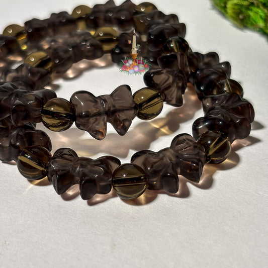 A close-up view of beaded stretch bracelets featuring carved ribbon-bow beads and round spacer beads in translucent ice obsidian . The dark brown, glassy crystal bracelets are layered on a plain white surface with a small floral candle logo above them and a hint of green moss in the top right corner.