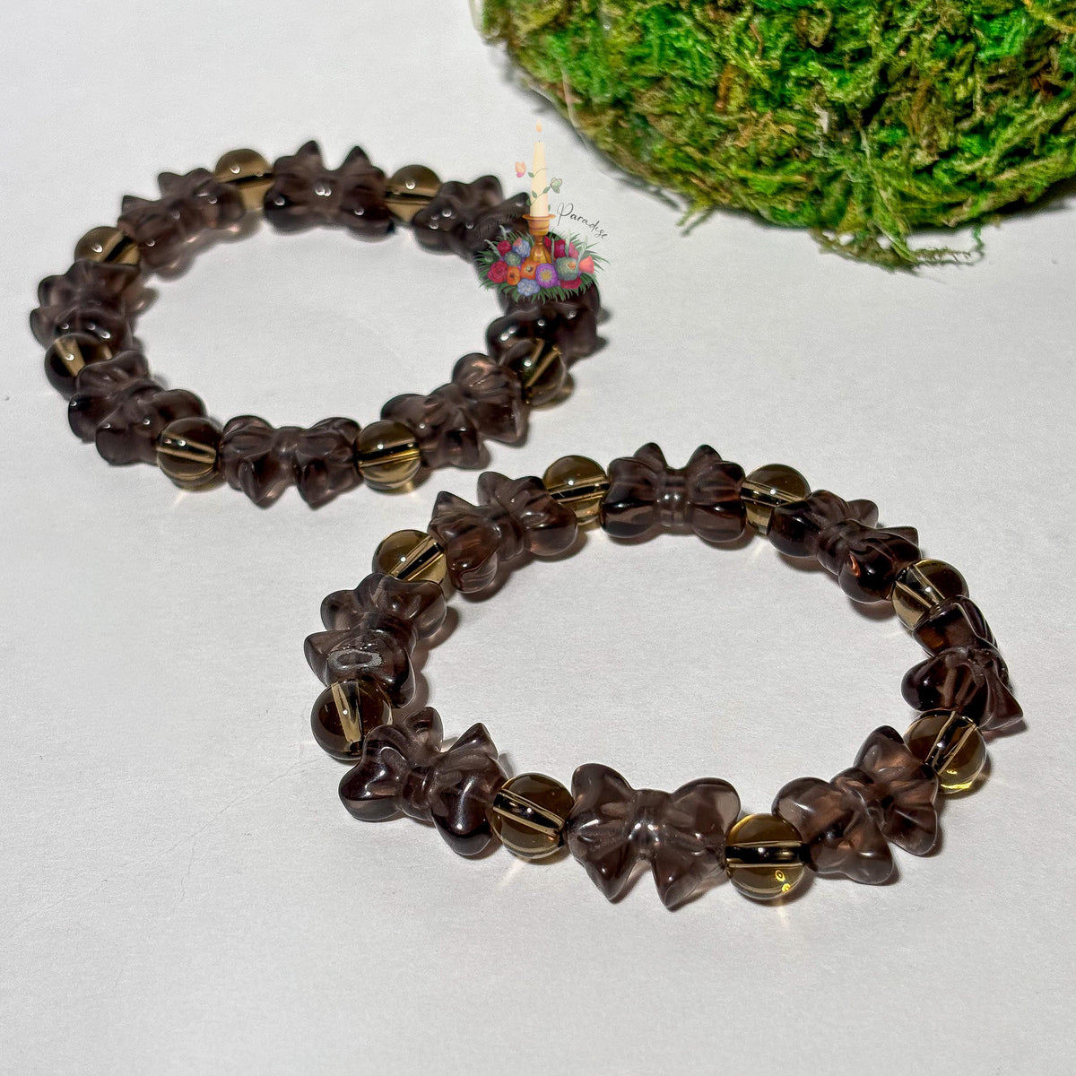 Two ice obsidian beaded stretch bracelets placed side by side on a smooth white surface, each featuring carved ribbon-bow beads alternating with polished round spacer beads in a translucent, smoky brown tone. Vibrant green moss fills the top right corner, and a small floral candle logo rests near the upper bracelet.