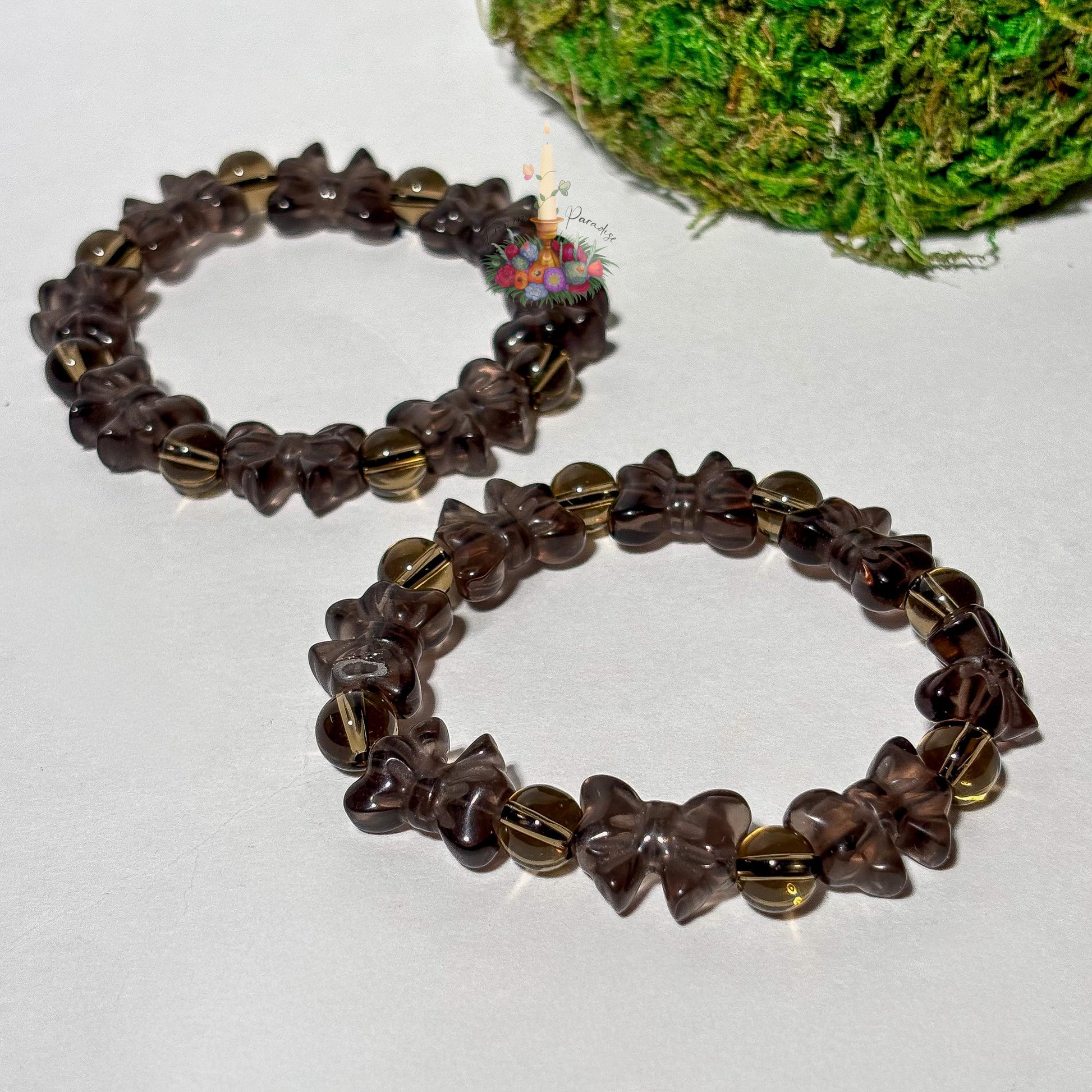 Two ice obsidian beaded stretch bracelets placed side by side on a smooth white surface, each featuring carved ribbon-bow beads alternating with polished round spacer beads in a translucent, smoky brown tone. Vibrant green moss fills the top right corner, and a small floral candle logo rests near the upper bracelet.