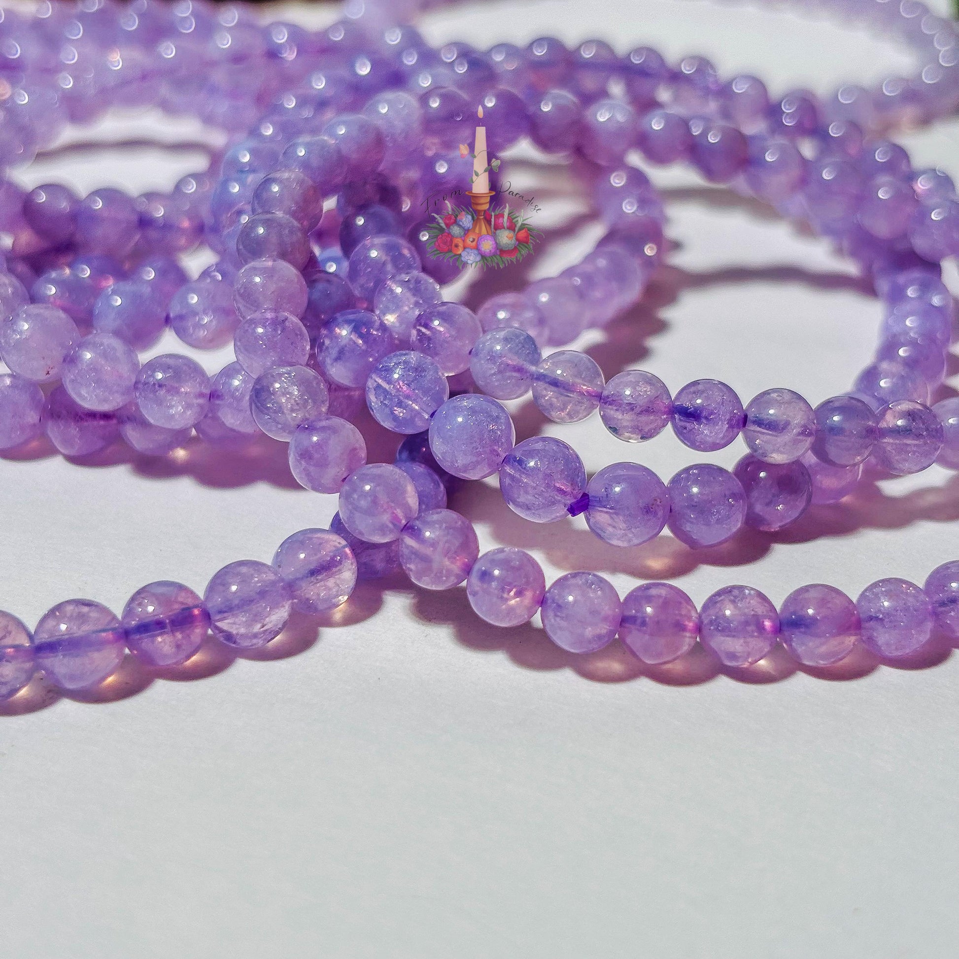 A cluster of round Lavender Moon Quartz beaded stretch bracelets piled together on a plain white surface. A carved stone statue sits out of focus in the background on the left, with vibrant green moss on the right and a floral candle logo centered above the bracelets.