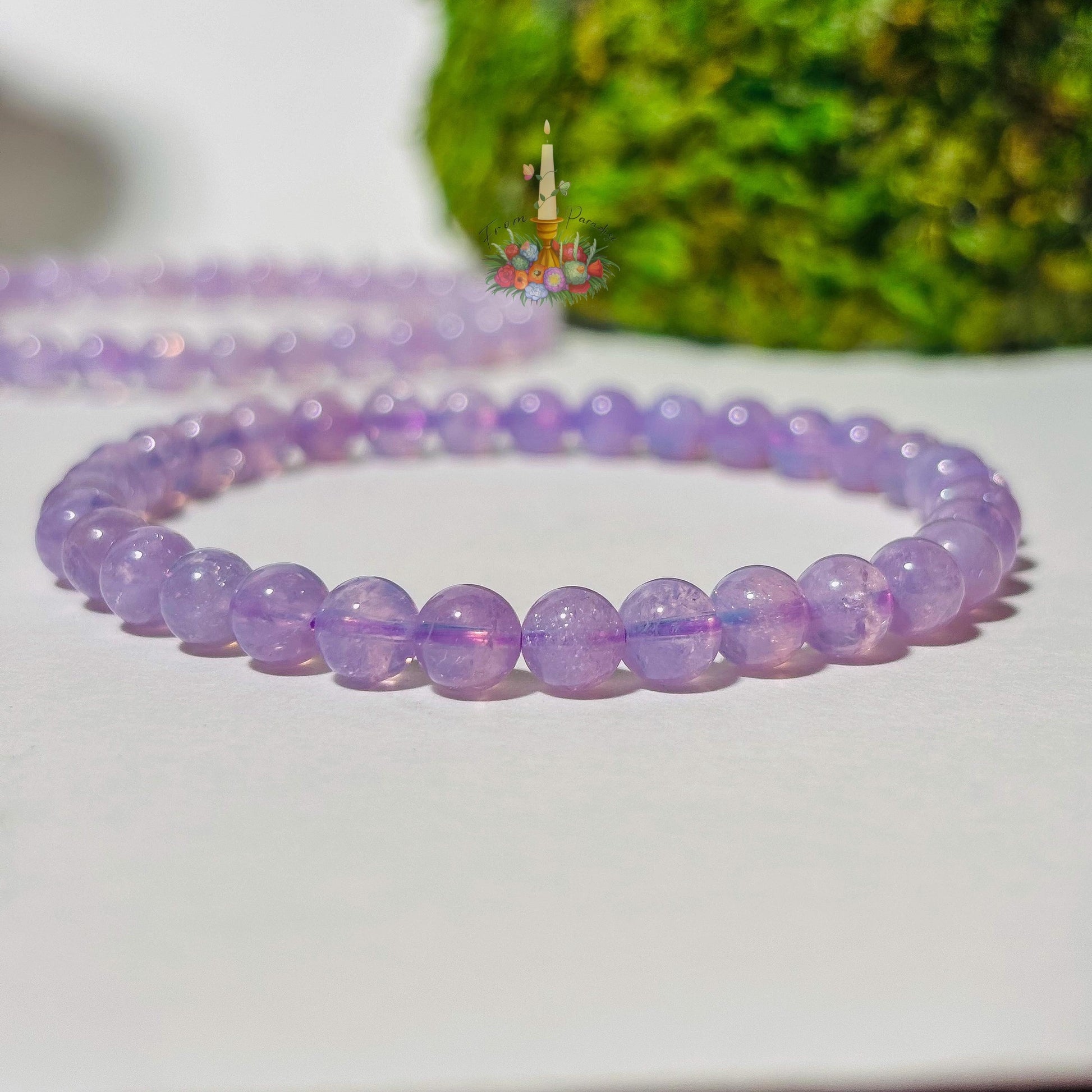A beaded stretch bracelet made of translucent, pastel lavender moon quartz polished round beads rests on a smooth white surface. In the background, a second matching beaded bracelet is softly blurred, alongside vibrant green moss and a floral candle logo.