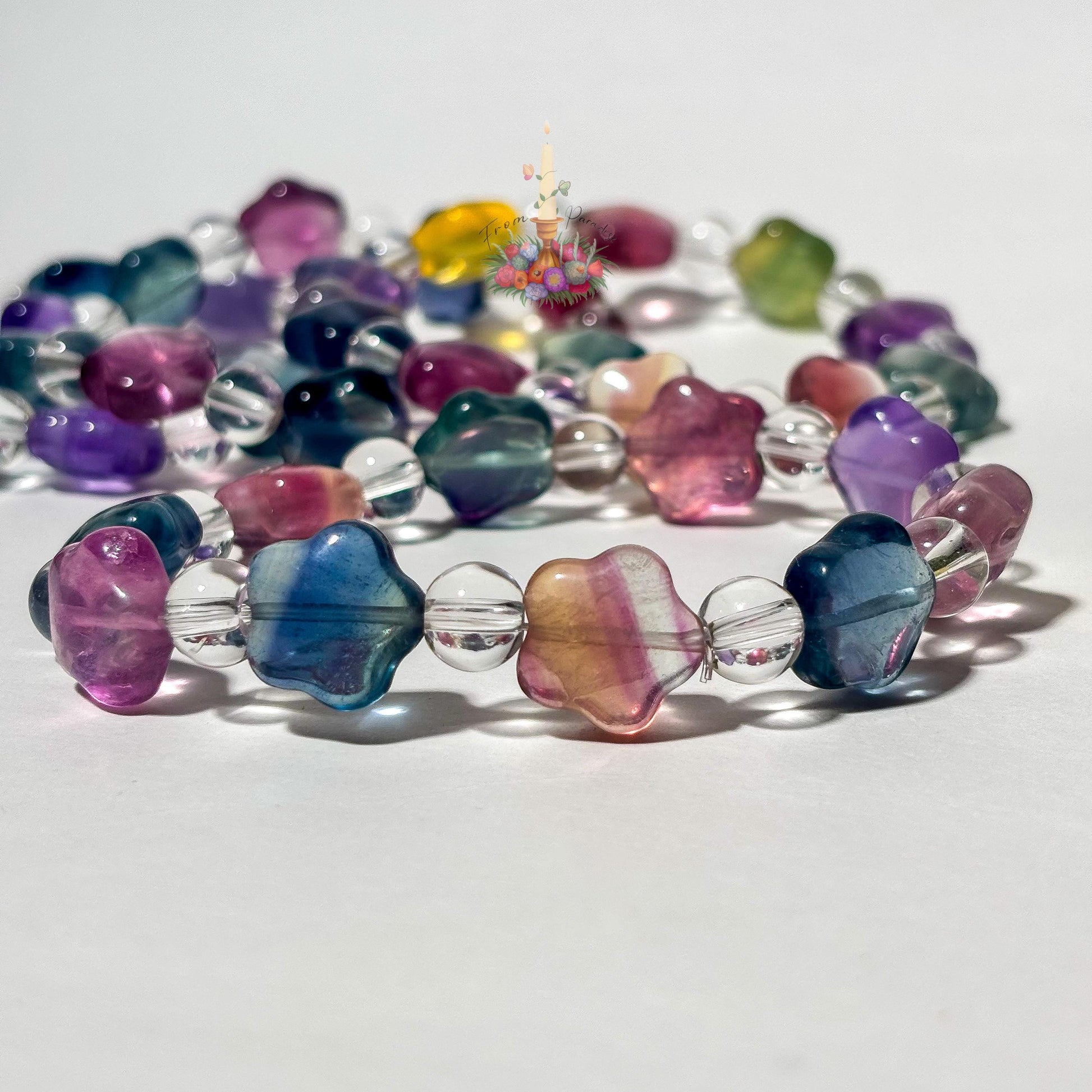 A close-up view of several beaded stretch bracelets on a white surface, with the foreground focusing on translucent, star-shaped fluorite beads in vibrant shades of magenta, blue, and yellow-purple color-zoning, separated by round clear quartz spacer beads. Additional matching fluorite and clear quartz bracelets lie softly out of focus in the background beneath a small floral candle logo.