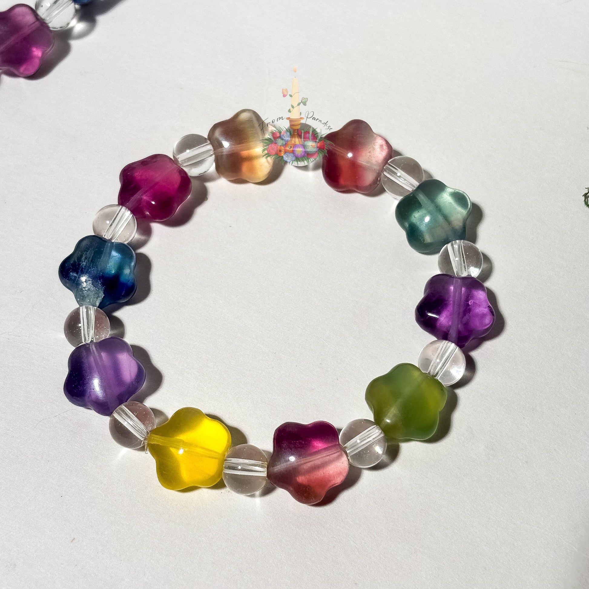 A single beaded stretch bracelet made of star-shaped fluorite beads in multi-colored pastel shades—including yellow, purple, deep pink, teal, and green—alternating with clear quartz round beads. The bracelet forms a circular layout against a smooth white surface, with a small floral candle logo positioned near the top center.