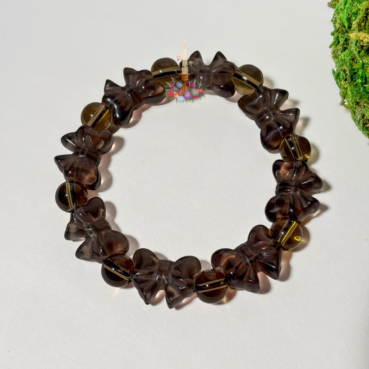 A single beaded stretch bracelet crafted from translucent ice obsidian, arranged in a circular loop against a clean white surface. The bracelet features carved ribbon-bow ice obsidian beads alternating with round spacer beads in the same smoky, translucent brown tone. A small floral candle logo sits near the top center of the loop, with a touch of green moss visible along the right edge.
