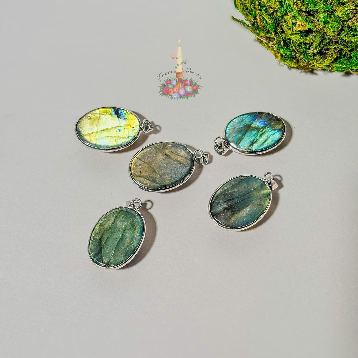 labradorite crystal pendants set in silver bezels are arranged on a plain white surface. Each pendant showcases a unique natural flash of color, ranging from bright yellow and blue to deep green and grey. A small patch of green moss is visible in the top right corner, and a small decorative logo with a candle and flowers sits in the upper center.
