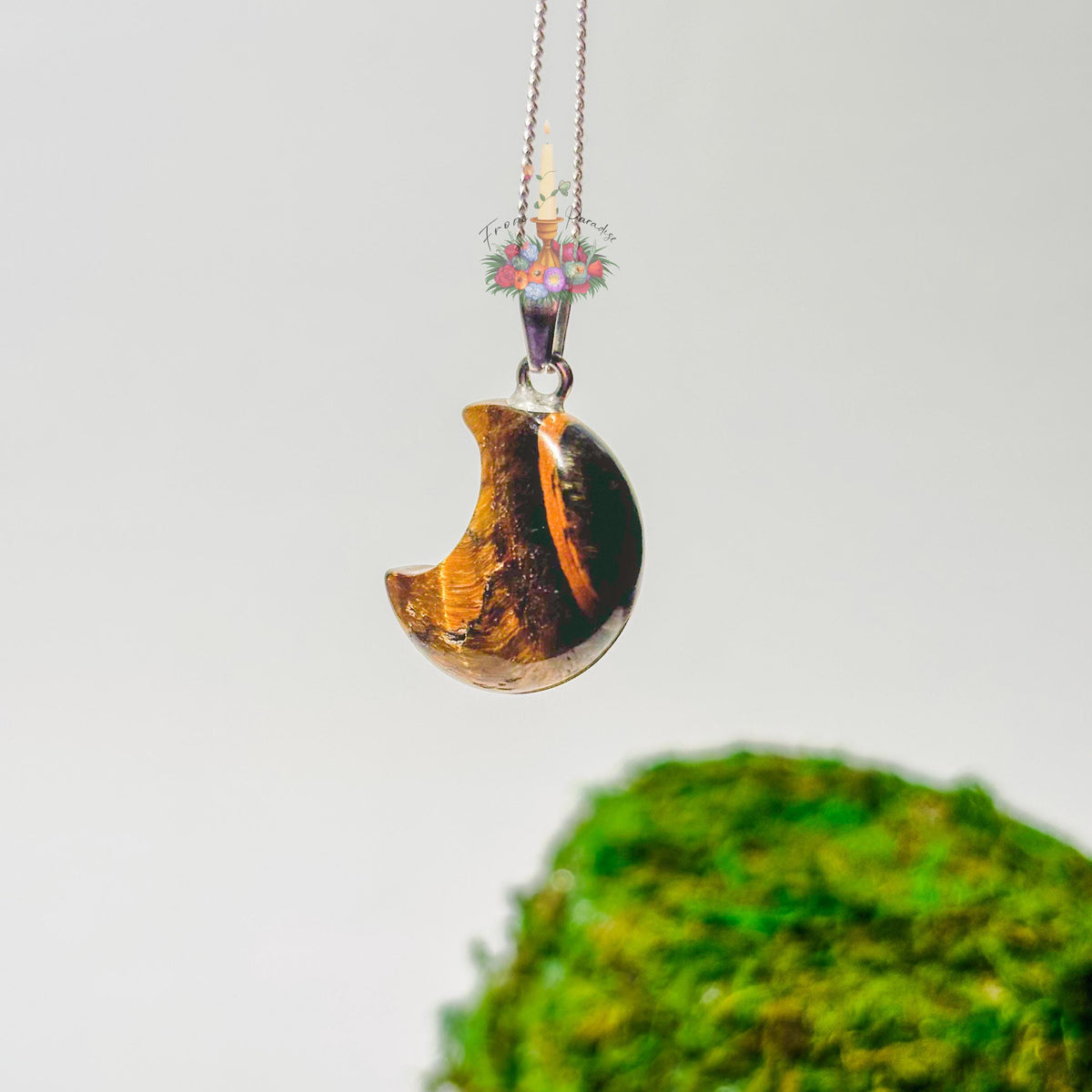 A crescent moon pendant necklace made of polished brown and orange wood with a silver-toned chain, shown against a light gray background with a blurred green mossy object at the bottom.